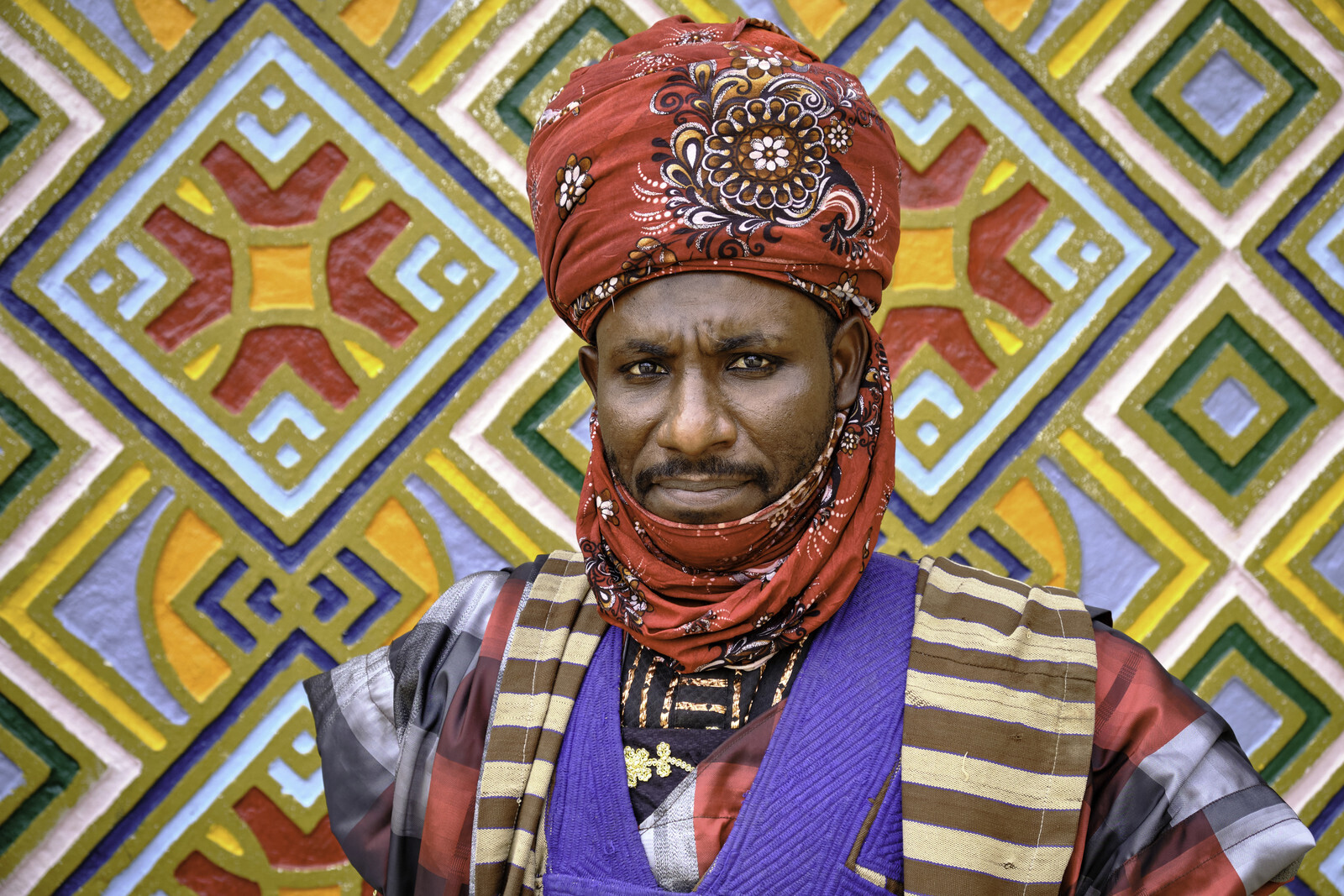 Servant of the Emir's palace in the city of Dutse dressed in a colorful traditional uniform next to a wall decorated with colorful Hausa motifs.