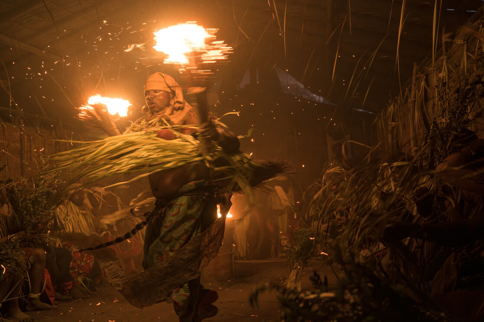 Participants in a Bwiti ceremony, dressed in ritual attire made from dried palm leaves, dance with lit torches inside a small temple. Bwiti is an animistic practice in which participants consume iboga, a plant that alters consciousness and induces visions, enabling them to communicate with the spirits.